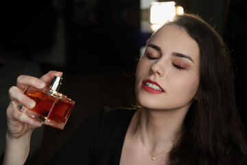 Woman with dark, wavy hair and red lipstick spraying perfume on her neck, eyes closed in a studio-like indoor setting with dramatic lighting