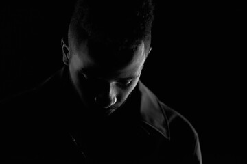 Black and white portrait of African American guy in leather jacket standing in the dark alone and looking down sadly