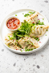 Steamed dumplings (momos) served on a white plate with a dipping sauce, garnished with fresh herbs. The dish is presented on a minimalist gray background, top view.