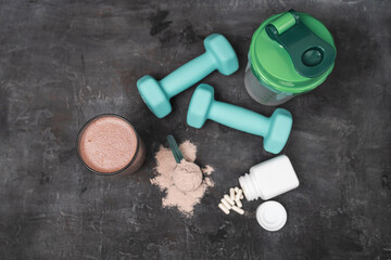 Top view of protein shake glass with whey protein powder scoop, white amino acid capsules, fitness dumbbells and shaker bottle on dark surface, sport nutrition and healthy lifestyle concept