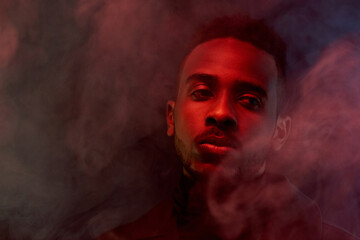 Portrait of attractive African American guy surrounded by smoke standing in the dark and looking...