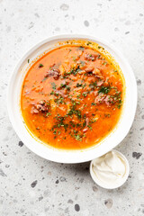 A top-down view of a hearty red soup garnished with fresh parsley in a white bowl, served with a side of sour cream on a speckled gray stone background.