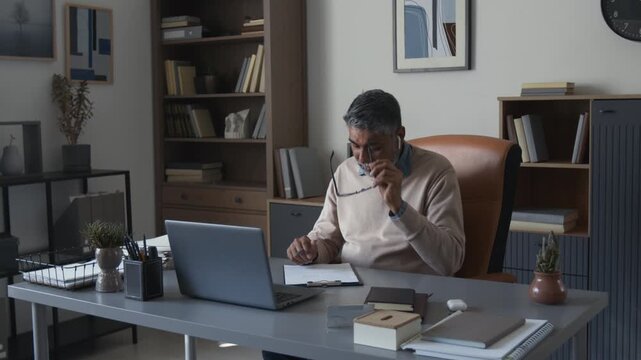Medium shot of mature Indian man as professional male therapist speaking in online therapy session with client via video call looking at laptop screen in office and taking notes, copy space