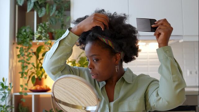 Morning routine of black woman styling natural curly hair with afro wide-tooth pick comb in front of mirror in cozy home with plants. Confident african american female gently detangling beauty curls.