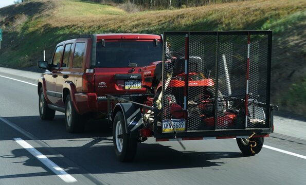 Pennsylvania, U.S - Oct 18, 2025 - A red Jeep Commander towing an open utility trailer loaded with red lawn maintenance equipment on a highway.