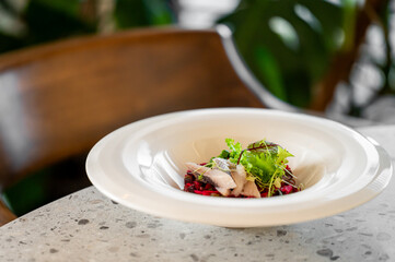 A close-up of a gourmet herring and beetroot salad on a white plate, garnished with fresh greens. The dish is placed on a speckled stone countertop in a restaurant setting.