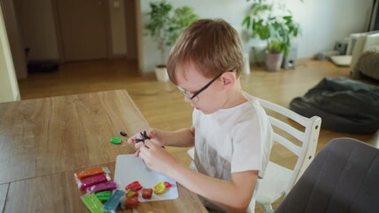 Boy with glasses sitting at wooden table molding colorful clay pieces into shapes with focus and creativity, engaging in artistic activity, developing imagination at home - Powered by Adobe