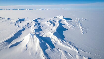 An aerial view of a snowy, mountainous landscape under a bright blue sky with a distant, foggy horizon, casting shadows