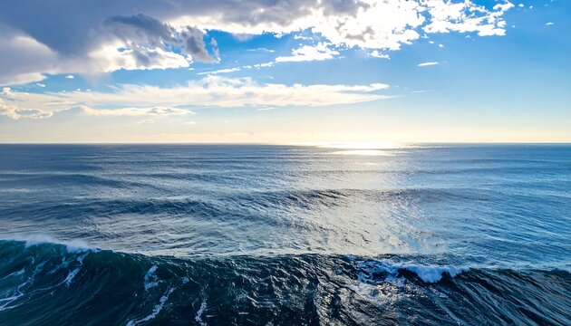 Aerial view captures a vast, serene ocean under a partly cloudy, radiant sky. Gentle waves roll. Sunlight sparkles on water's surface