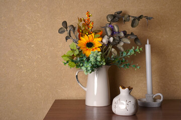 Rustic floral arrangement in a white jug on a wooden shelf next to a tall candle and decorative ceramic item. The bright sunflower and eucalyptus leaves create a cozy atmosphere.