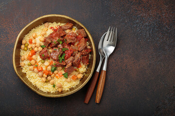 Traditional dish of Maghreb cuisine, a Moroccan couscous with lamb or beef, vegetables, chickpeas, and peas in a rustic bowl with cutlery on dark background, copy space