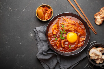 Top view of spicy kimchi jjigae soup in a black bowl with egg yolk, enoki, shiitake, tofu and green onions, with chopsticks and cooking ingredients, cuisine of Korea, copy space