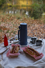 An autumn picnic in the park. A thermos, tin mugs, and hot dogs sit on a blanket on a wooden table.