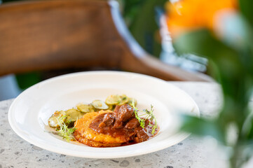 Braised meat with a rich brown sauce served on crispy potato pancakes, garnished with fresh herbs and sliced pickles, on a white plate on a speckled table.