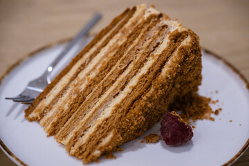 A slice of layered honey cake with cream filling sits on a white-rimmed plate beside a raspberry and scattered crumbs.