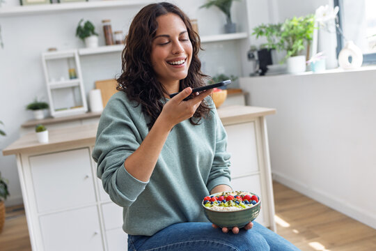 Pretty woman eating a healthy fruit bowl while taking a photo with her smartphone in the kitchen at home