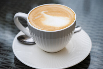 A freshly made cappuccino with delicate milk froth sits on a white saucer atop a woven café table.