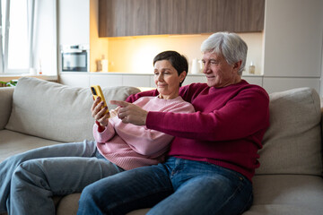 Engaged senior spouses lean together captivated by digital content on smartphone. Web feed surfing, social media on retirement, active seniors leisure time with phone, happy joyful family rest on sofa
