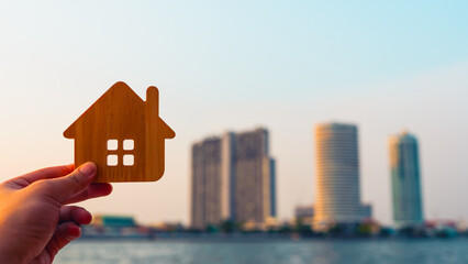 Hand holding wooden house icon with blurred city skyline background, symbolizing real estate, urban living, property investment, or housing business concepts.
