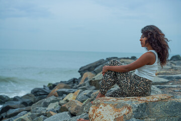 A woman sits on a rock by the ocean, looking out at the water