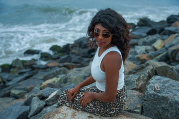 A woman with curly hair and glasses is sitting on a rock by the ocean