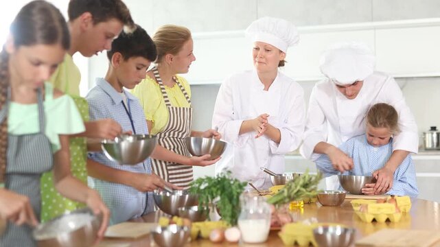 Two experienced chefs, friendly woman and man in white uniform giving culinary lesson to group of inquisitive tweens, teaching basic cooking skills
