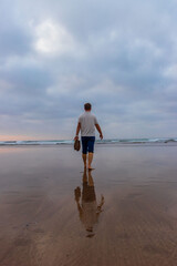 Back view of man walking barefoot on beach with shoes in hand, calm sea and reflection on wet sand, concept of travel, freedom, movement and solitude.