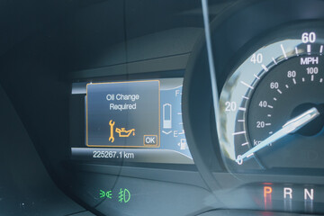 Close-up of a modern car dashboard, displaying an Oil Change Required warning light and message. Digital gauge cluster showing speedometer, fuel and battery status with high mileage