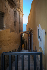 Narrow downhill lane with stairs between cliffside houses in Positano, Italy, with a sliver of blue sky and sea visible in the distance