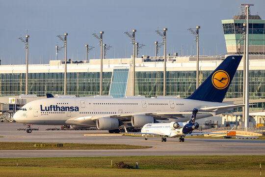 Ein Airbus A380-800 der Lufthansa steht am Terminal des Flughafens M&uuml;nchen und bereitet sich auf den Abflug . Im Vordergrund rollt ein Bombardier CRJ-900 der Lufthansa CityLine 