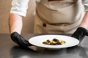 A chef wearing a white jacket and black gloves presents a perfectly plated gourmet dish on a white plate. The focus is on the artfully arranged food and the hands.