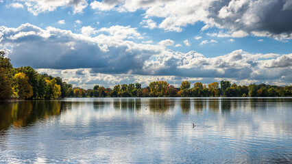 Obraz premium Blick über den Dutzendteich in Nürnberg, Bayern, mit herbstlich gefärbten Bäumen am Ufer und dramatischer Wolkenformation am Himmel. 