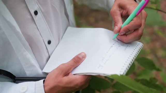 Hand view of researcher in white coat writing notes on jotter with pen in forest, documenting ecological observations during field research surrounded by leaves and natural environment