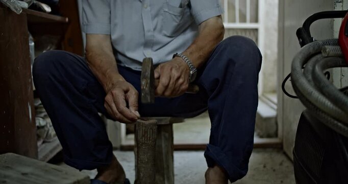 Witness the meticulous work of an elderly craftsman as his weathered hands expertly hammer a small metal piece on a sturdy wooden stool.