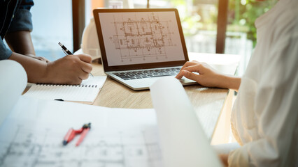 A man and a woman are working on a laptop together. The man is writing on a piece of paper while the woman is typing on the laptop. The laptop screen shows a drawing of a building