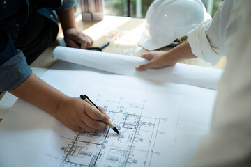 Two people are working on a blueprint of a building. One of them is holding a pen and drawing on the paper. The other person is wearing a hard hat