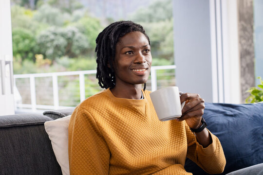 African American man in mustard sweatshirt sitting on sofa at home holding mug checking smartwatch