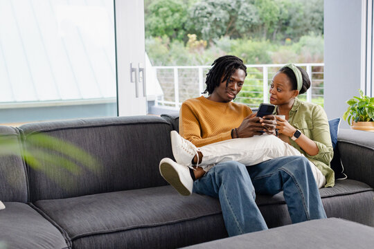 African American couple sitting on grey sofa in living room, holding smartphone and ceramic mug - Powered by Adobe