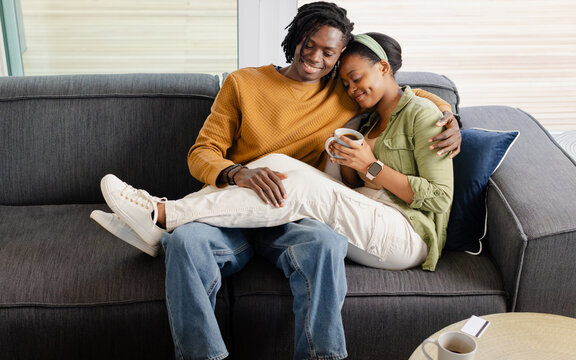 African American couple cuddling on grey couch in home living room holding mug wearing smartwatch