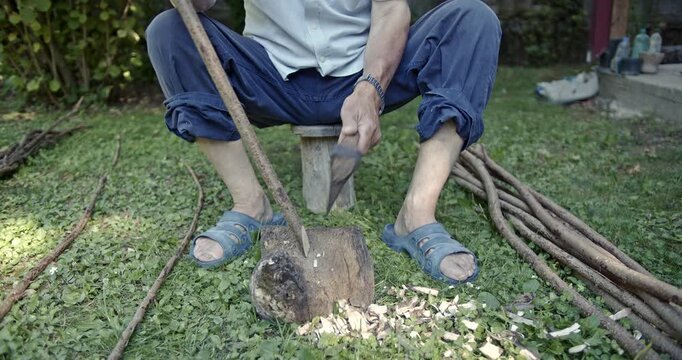 Close-up of an experienced man's hands as he expertly uses a small axe to strip bark from a long wooden stick in a natural outdoor setting, showcasing traditional craftsmanship and manual labor.