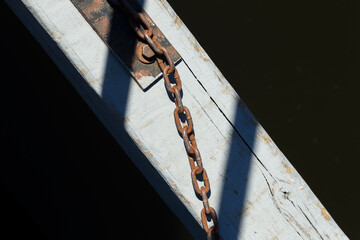 Rusty chain attached to weathered wooden beam over dark water. Old metal chain and shadow detail on sunlit dock surface.