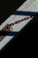 Rusty chain attached to weathered wooden beam over dark water. Old metal chain and shadow detail on sunlit dock surface.
