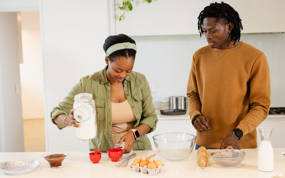 African American couple pouring flour into red measuring cups and sifting cocoa at kitchen island