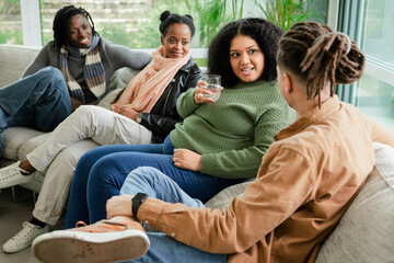 African American friends sitting on living room sofa by window, holding water glass while chatting