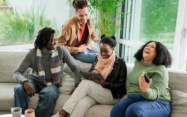Diverse friends sharing tablet and using smartphone on gray sofa in bright living room