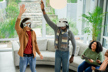 Diverse friends standing in living room wearing VR headsets raising arms with friend using tablet