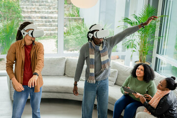 Diverse friends wearing VR headsets and exploring virtual world near sofa in bright living room