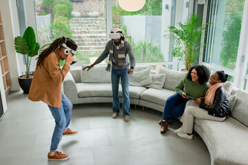 Diverse friends using VR headsets and laughing holding game controllers on gray sofa in living room