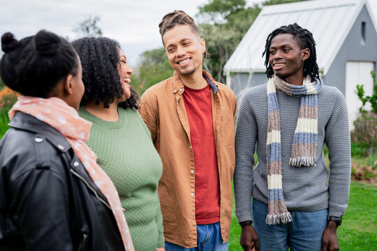 Diverse friends chatting wearing leather jacket sweater scarves in grassy field at modern cabins