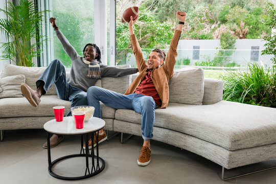 Diverse male friends cheering on curved sofa in modern living room with football, popcorn, red cups
